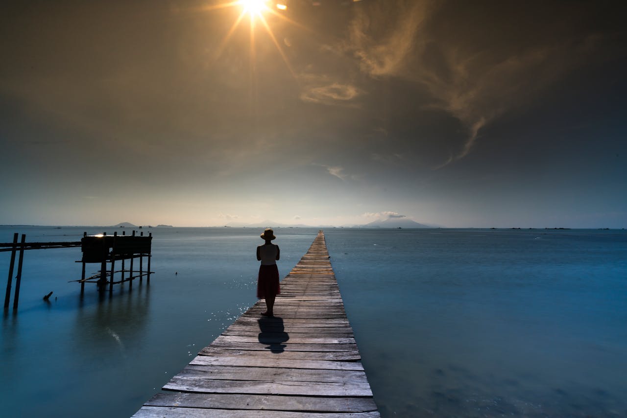 A person in silhouette walks on a sun-drenched boardwalk over a tranquil ocean.