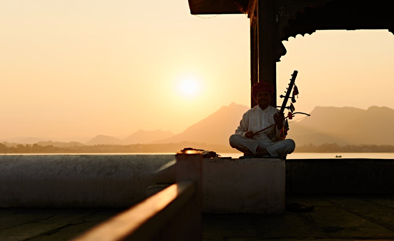 A musician playing ravanatha at sunset by a serene lake, dressed in traditional attire.
